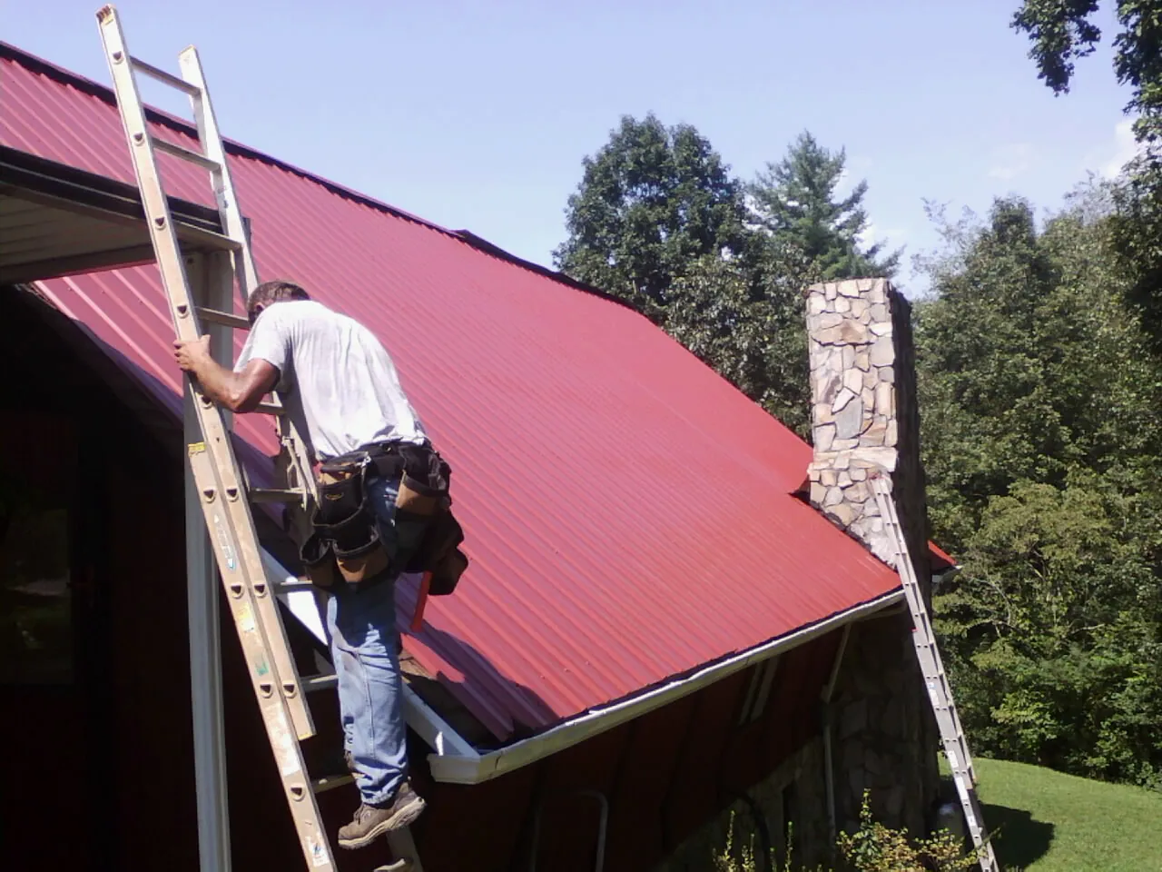 Roofer on a ladder beside a red metal roof and stone chimney — Soffit & Fascia Repair by Roofing Brothers in Warrensville